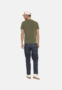 T-shirt vert olive, jeans bleu foncé avec accent vert sur la poche, baskets blanches avec semelle beige, et casquette blanche avec détail orange.