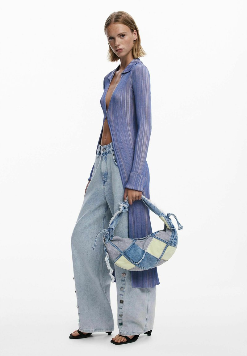 Donna che indossa un lungo cardigan trasparente viola, jeans in denim chiaro a gamba larga con dettagli in bottone, sandali neri e tiene una borsa in denim patchwork.