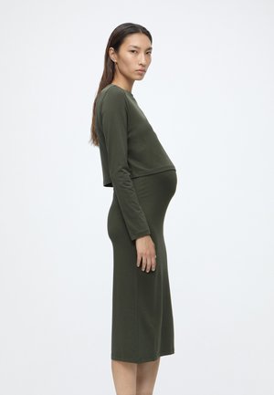 Femme enceinte portant un top moulant à manches longues et une jupe vert olive, debout de profil devant un fond clair uni.