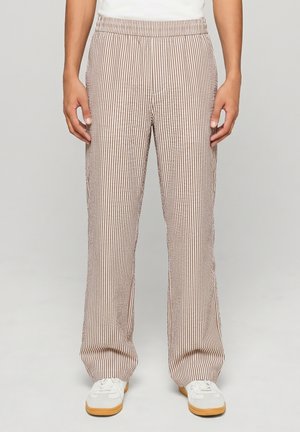 Personne portant un pantalon ample à rayures verticales beiges et blanches et des baskets blanches avec semelles en gomme, debout devant un fond clair uni.