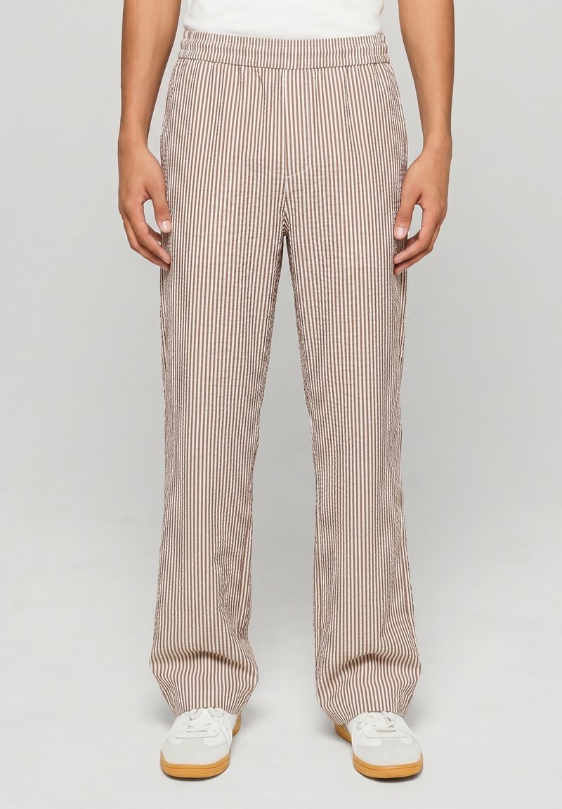 Personne portant un pantalon ample à rayures verticales beiges et blanches et des baskets blanches avec semelles en gomme, debout devant un fond clair uni.