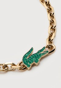 Βραχιόλι αλυσίδα από χρυσό με γοργόνα κοσμημένη με πράσινες πέτρες, συνδεδεμένη ανάμεσα σε χοντρούς κρίκους αλυσίδας σε λευκό φόντο.