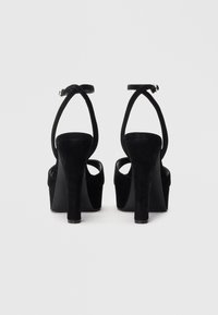 Zwarte suède plateauhakken met enkelbandjes en blokhakken, van achteren getoond op een witte achtergrond.