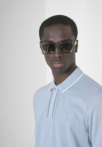 Polo bleu clair en tissu texturé, avec des accents de rayures subtiles sur le col et un logo au centre. Lunettes de soleil rectangulaires foncées.