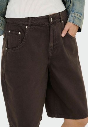 Personne portant un short en denim marron foncé à coupe ample avec des poches avant, associé à un haut blanc côtelé et une veste en denim, mains dans les poches.