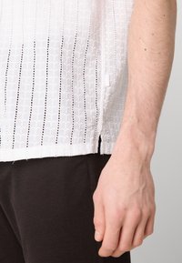 Witte blouse met een patroontextuur en verticale uitsparingen, gecombineerd met een donkerbruine stof, die een relaxed fit en zijsplitten laat zien.