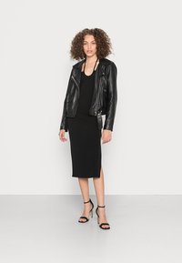 Jaqueta de motoqueiro preta em couro com lapelas, combinada com um vestido preto ajustado com uma fenda lateral. Modelo usa saltos altos pretos com tiras.