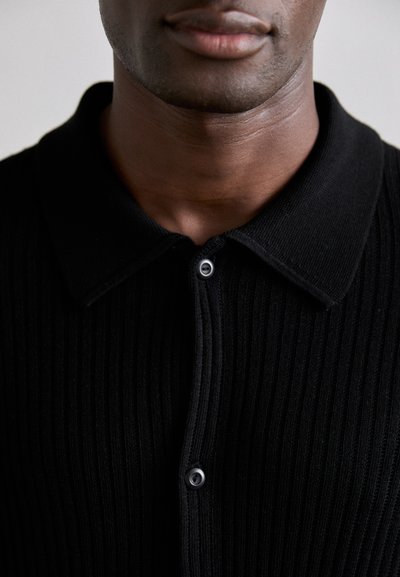Svart ribbad polo tröja med knäppning framtill och klassisk krage, tillverkad av ett strukturerat tyg med subtil lyster.