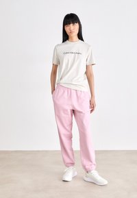 Lichtgrijs T-shirt met "Calvin Klein Jeans" logo, gecombineerd met roze joggers met elastische tailleband en witte sneakers, tegen een effen achtergrond.