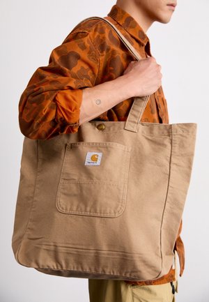 Osoba nosząca pomarańczową koszulę moro, niosąca dużą beżową torbę Carhartt na ramieniu, pokazująca tatuaż na przedramieniu oraz szczegóły kieszeni torby.