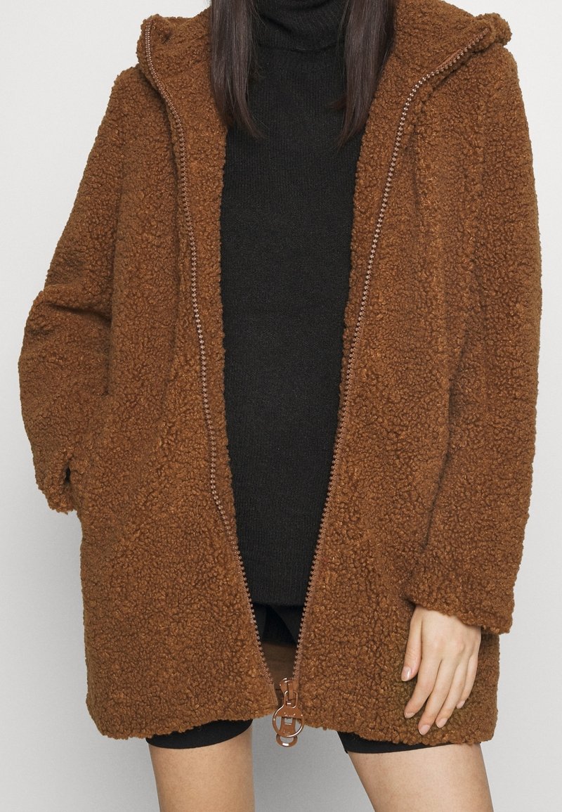 Femme portant un manteau teddy marron oversize avec une grande fermeture éclair sur un col roulé noir et un short noir, sur fond uni.
