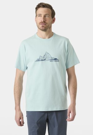 Hombre con camiseta azul claro de Helly Hansen con gráfico de montaña y pantalones oscuros, de pie contra un fondo claro y liso.