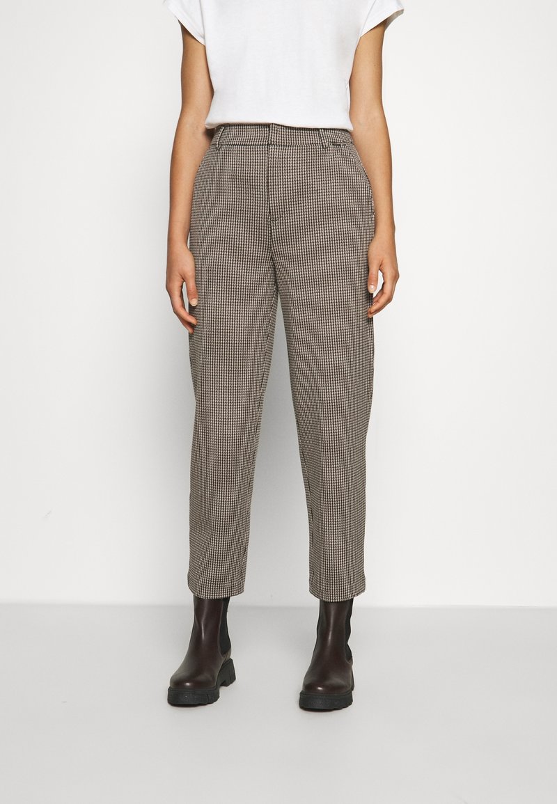 Femme portant un pantalon marron taille haute à motif pied-de-poule, un haut blanc à manches courtes et des bottes noires épaisses, debout devant un fond blanc uni.