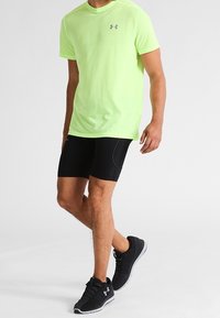Camisa de manga corta amarilla neón con logo gris, combinada con pantalones cortos ajustados negros y zapatillas deportivas negras. Diseño sencillo, tejido ligero.