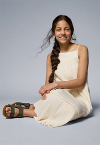 Light beige sleeveless dress with a textured fabric and a straight neckline. Green sandals with two straps and a cork footbed.