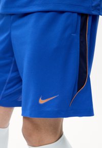 Μπλε αθλητικό σορτς με πορτοκαλί λογότυπο Nike Swoosh και μαύρες πλαϊνές πινελιές με πορτοκαλί περιθώριο, φορεμένα με λευκές κάλτσες.