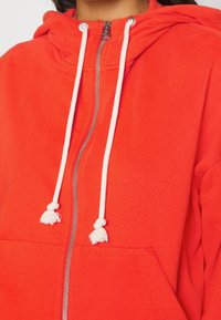 Sudadera con capucha roja y cremallera completa, con cordones blancos y bolsillo tipo canguro. La tela es suave y tiene una textura lisa.