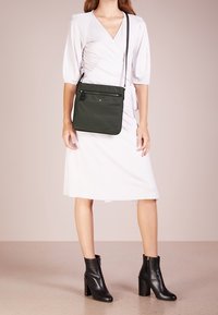 Mørkegrøn rummelig skuldertaske med glat tekstur, der har en lynlåslomme og justerbar rem, båret over en lysefarvet omslagskjole.