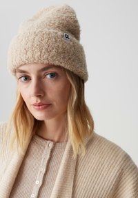Berretto a maglia beige e morbido, con la parte superiore piegata e una piccola etichetta nera; indossato con un cardigan beige a coste abbinato e uno sfondo morbido.