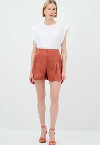 Camicia bianca a maniche corte con punti testurizzati, abbinata a pantaloni corti a vita alta di colore ruggine con pieghe. Modella indossa sandali con cinturini abbinati.