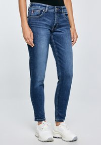 Nő karcsúsított kék farmerben és fehér tornacipőben, egyszínű világos háttér előtt állva.