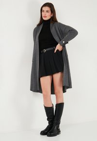 Cardigan gris à côtes avec manches retroussées, col roulé noir, jupe plissée noire et bottes noires à hauteur de genoux avec accents dorés.