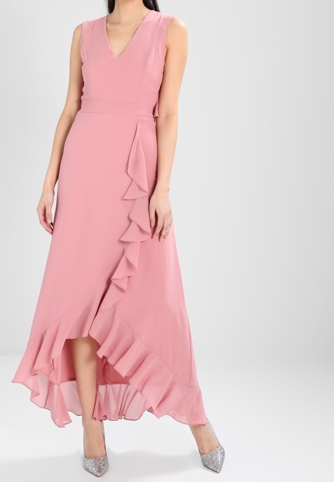 Femme portant une robe rose sans manches avec un ourlet asymétrique à volants et des talons hauts argentés pailletés, debout devant un fond uni.