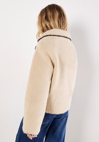 Beige fleecedeken met een kraag, zwarte gestikte accenten en een rechte pasvorm. De manchetten en de zoom hebben een zigzagpatroon.