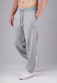 Man wearing light gray sweatpants with drawstring, white low-top sneakers, and a silver bracelet, standing against a plain background.
