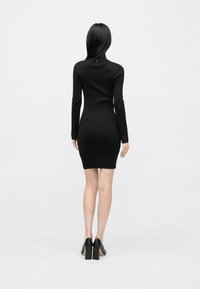 Robe noire moulante à manches longues, col montant et poignets texturés. Texture lisse, silhouette ajustée et ourlet à hauteur du genou. Portée avec des talons noirs.