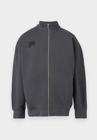 Veste zippée gris foncé avec col haut et petit logo noir brodé sur le côté gauche de la poitrine, présentée sur un fond clair.
