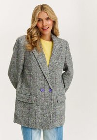 Žena s dlouhými blond vlasy, oblečená v šedém kostkovaném blejzru s fialovými knoflíky, pod kterým má na sobě žlutý svetr a světlé modré džíny.