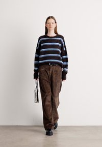 Gestreifter Pullover in Dunkelbraun mit blauen Streifen, kombiniert mit lockeren braunen Cordhosen. Das Modell hält eine metallische Handtasche und trägt schwarze Schuhe.