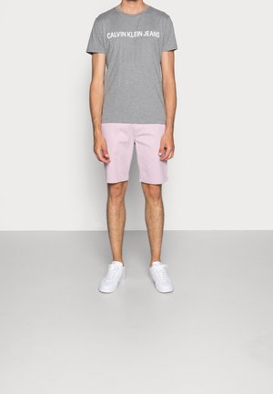 Mężczyzna ubrany w szarą koszulkę Calvin Klein Jeans, jasnoróżowe spodenki i białe trampki, stoi na jednolitym jasnym tle.