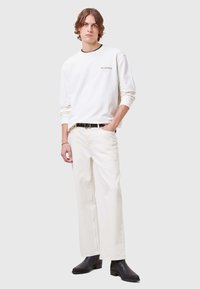 Felpa bianca con collo nero e logo, abbinata a pantaloni a gamba larga beige chiaro e stivali neri alla caviglia, con un design elegante.