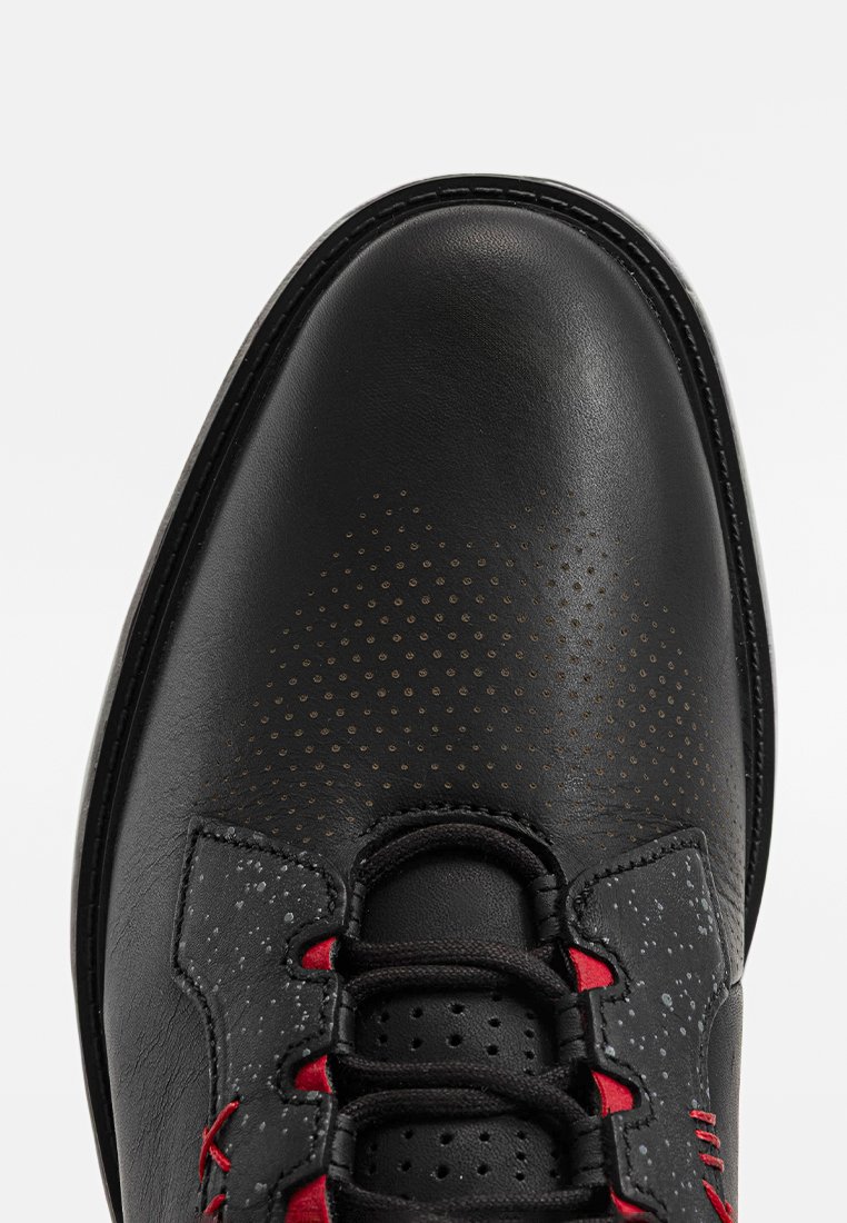Chaussure en cuir noir avec une tige texturée, présentant des détails perforés, des accents rouges sur les lacets et une conception à bout arrondi.