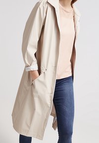 Béžový voděodolný trenčkot s volným střihem, zapínáním na knoflíky, ohrnovacími rukávy a bočními kapsami, vrstvený přes světle růžový top.