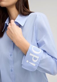 Camicia button-up azzurro chiaro con colletto. Un polsino presenta un testo stampato bianco. Il tessuto sembra liscio e leggero.