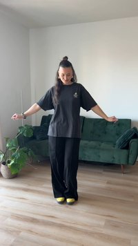 Μαύρο υπερμεγέθες T-shirt με λογότυπο, σε συνδυασμό με φαρδιά μαύρη παντελόνια και κίτρινα παπούτσια. Το άτομο στέκεται σε ένα φωτεινό δωμάτιο με πράσινο καναπέ.