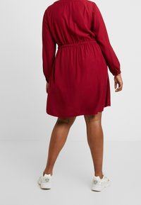 Abito rosso con maniche lunghe, vita arricciata, vestibilità morbida e scollatura rotonda. Abbinato a sneaker bianche, design minimalista.