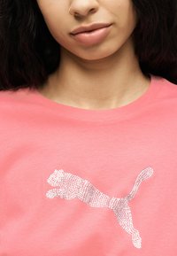 Κοντινό πλάνο ενός ατόμου που φοράει ροζ πουκάμισο με ασημένιο λογότυπο Puma από στρας στο στήθος.