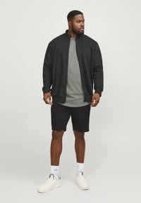 Black zip-up jacket over a green shirt, paired with black shorts and white sneakers. Features include simple design and athletic fit.