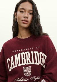 Ung kvinde med langt mørkt hår iført en kastanjebrun "University of Cambridge Athletic Dept." sweatshirt, som poserer mod en ensfarvet lys baggrund.