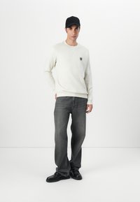 En ung mann står mot en hvit bakgrunn, iført en kremfarget genser, svart caps, grå jeans og svarte sko, i en avslappet posisjon.