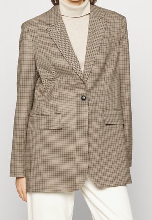 Kobieta ubrana w brązowo-beżową jednorzędową marynarkę w kratę, na kremowym golfie i białych spodniach, stojąca na tle jednolitego tła.