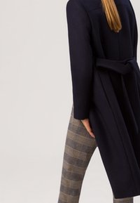 Cappotto lungo blu navy con vita aderente, abbinato a pantaloni fitted a quadri grigi. Il cappotto presenta un design snello e una texture liscia.