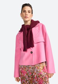Chaqueta ajustada de mezcla de lana rosa con dos bolsillos frontales, grandes solapas y un botón negro. Una bufanda de punto burdeos está drapeada alrededor del cuello.