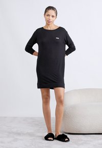 Black long-sleeve dress with a relaxed fit, rounded neckline, and small embroidered logo. Paired with black fuzzy slides.
