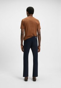 Bruine korte mouwen shirt en donkerblauwe op maat gemaakte broek met rechte pijpen, gedragen met donkere leren schoenen, gezien vanuit de achterzijde.