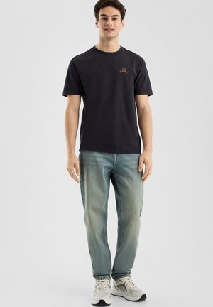 Zwarte katoenen t-shirt met korte mouwen en een ronde hals, voorzien van een klein gouden geborduurd logo. Gepaarde met lichtblauwe denim jeans.
